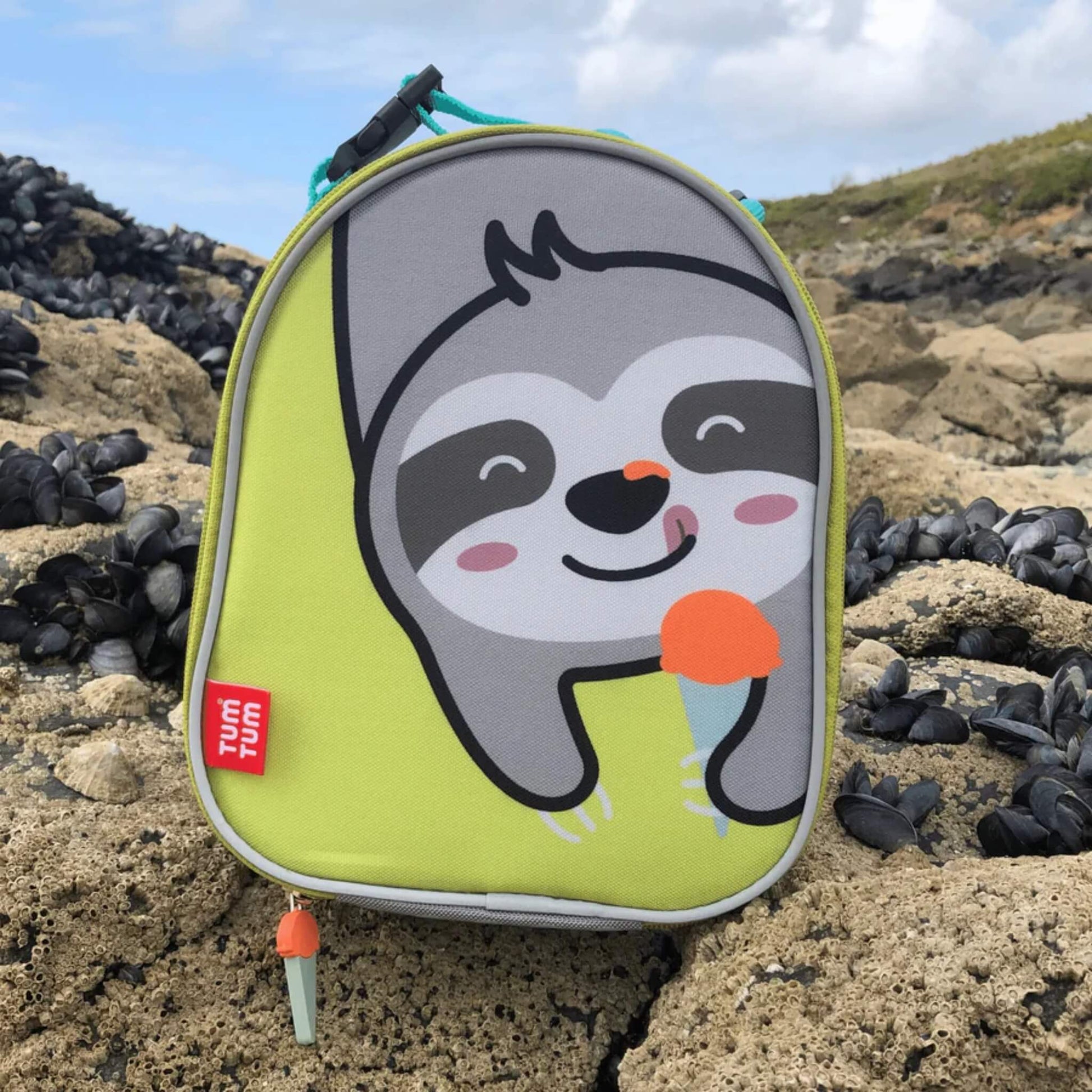 A lunch bag standing on rocks at the beach, showing the sloth character on the front with grey piping and a zip pull visible at the base.