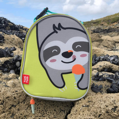 A lunch bag standing on rocks at the beach, showing the sloth character on the front with grey piping and a zip pull visible at the base.