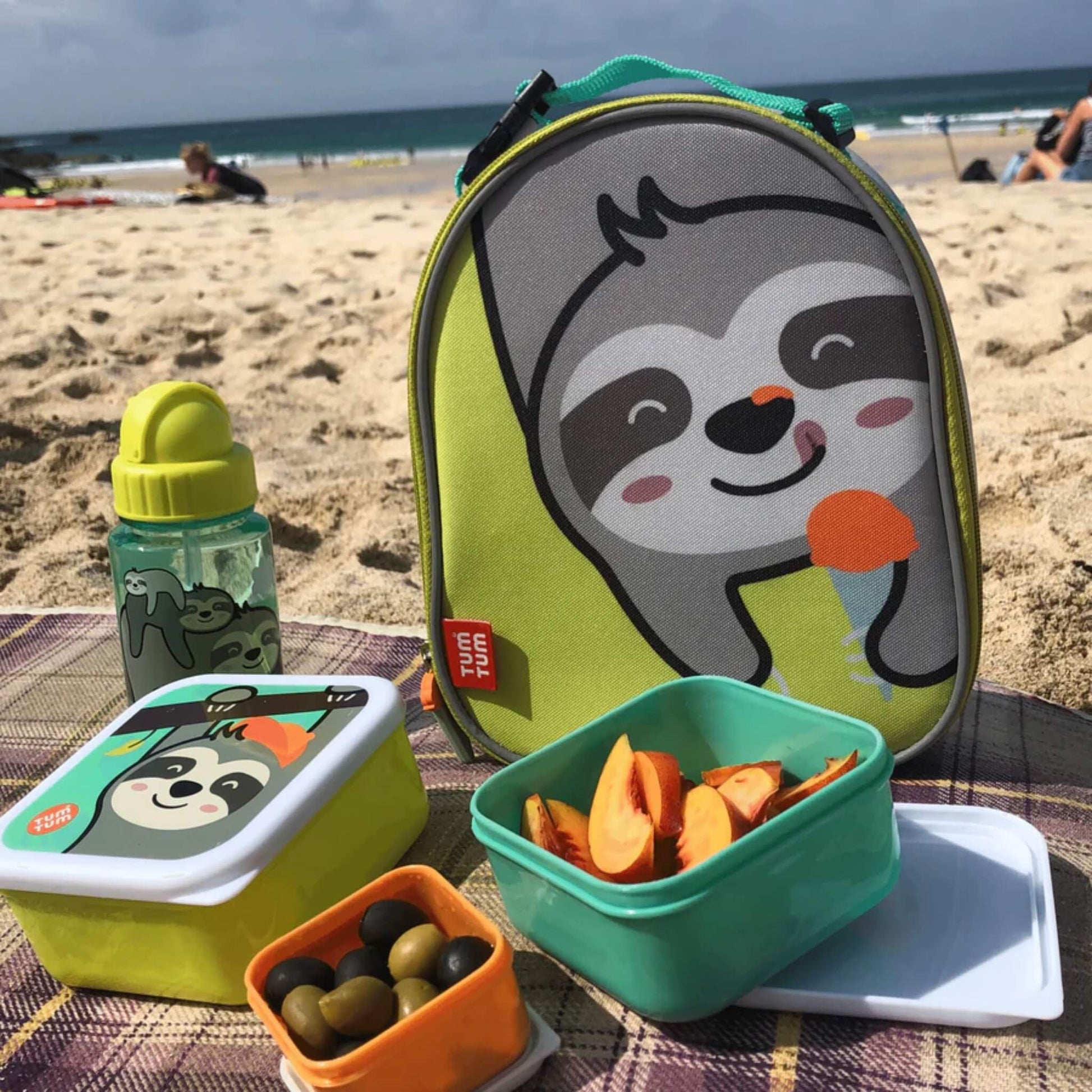 A picnic setup on sand with an insulated lunch bag behind several open food containers holding fruit, olives, and snacks, alongside a matching drinks bottle.