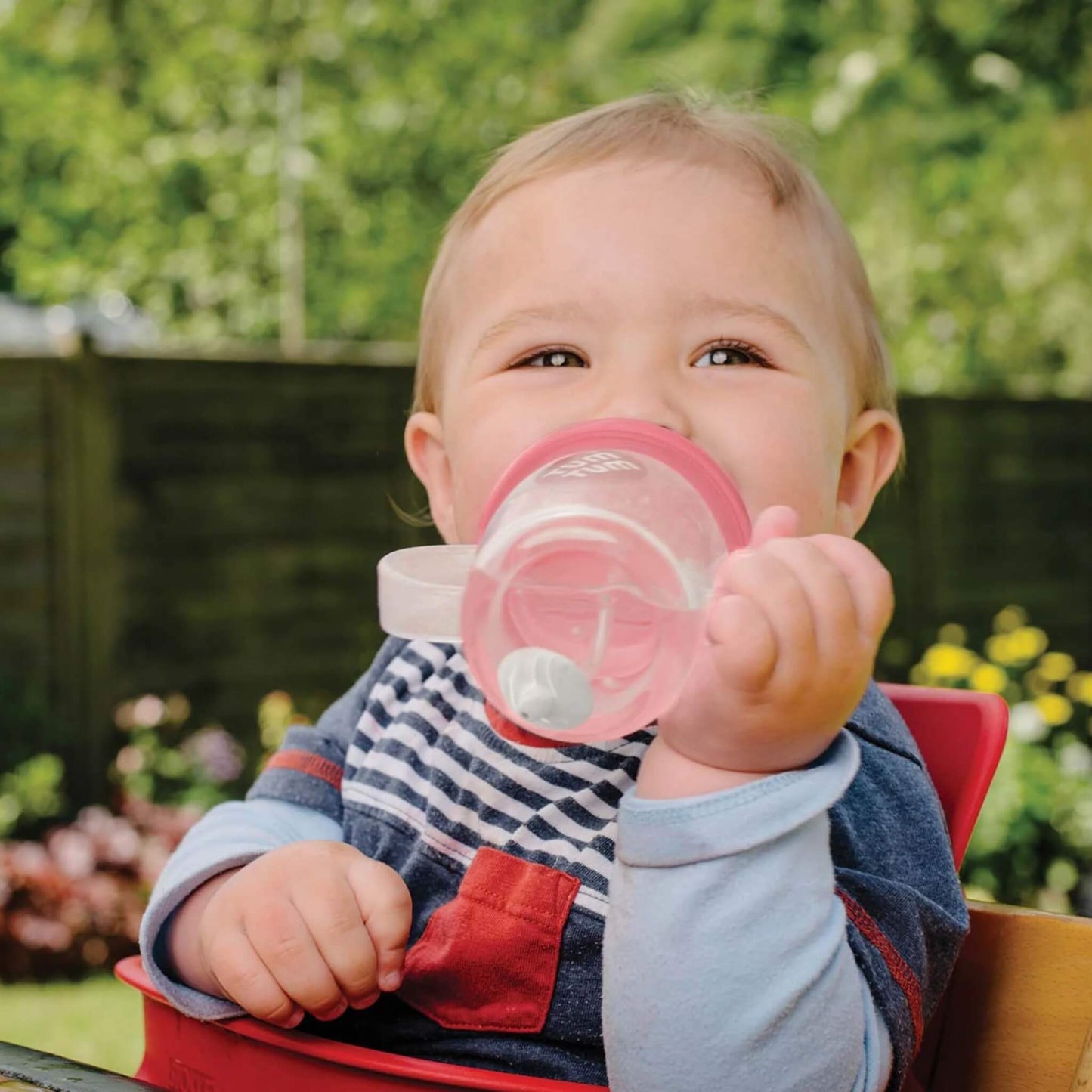 A young child seated outdoors drinking from a straw sippy cup, holding the cup independently with one hand.