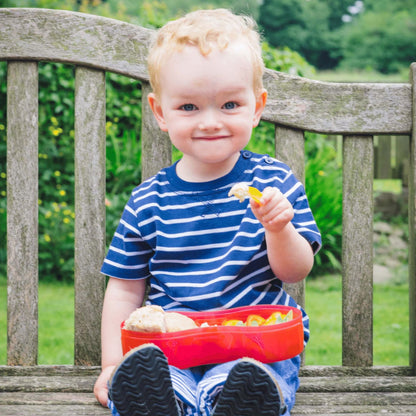 Young child enjoying a meal outdoors with a red lunch box and integrated dipping pot separator, perfect for healthy snacks on the go.