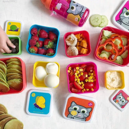 A colourful children's lunch set arranged on a white surface, including square plastic containers with cartoon-themed lids, a pink cat cup, sliced cucumbers, strawberries, crackers, hard-boiled eggs, corn and tomato pieces, and dip. A partial hand is visible at the left edge.