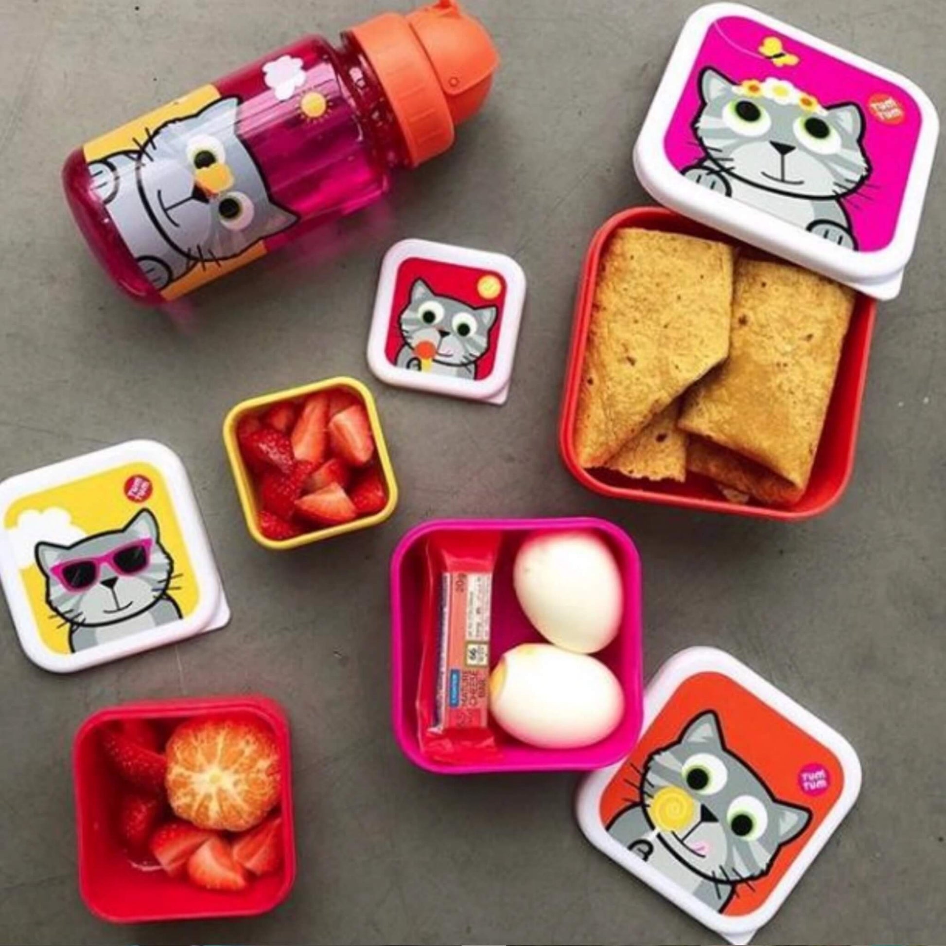 A set of cartoon cat-themed lunch containers and a matching bottle arranged on a grey surface, filled with snacks including crackers, strawberries, an orange slice, and hard-boiled eggs.