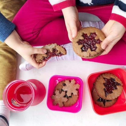 A top-down lifestyle shot of a person holding star-shaped cookies filled with dark red jam, with two more cookies in pink and red square dishes and a pink drink on a white surface.