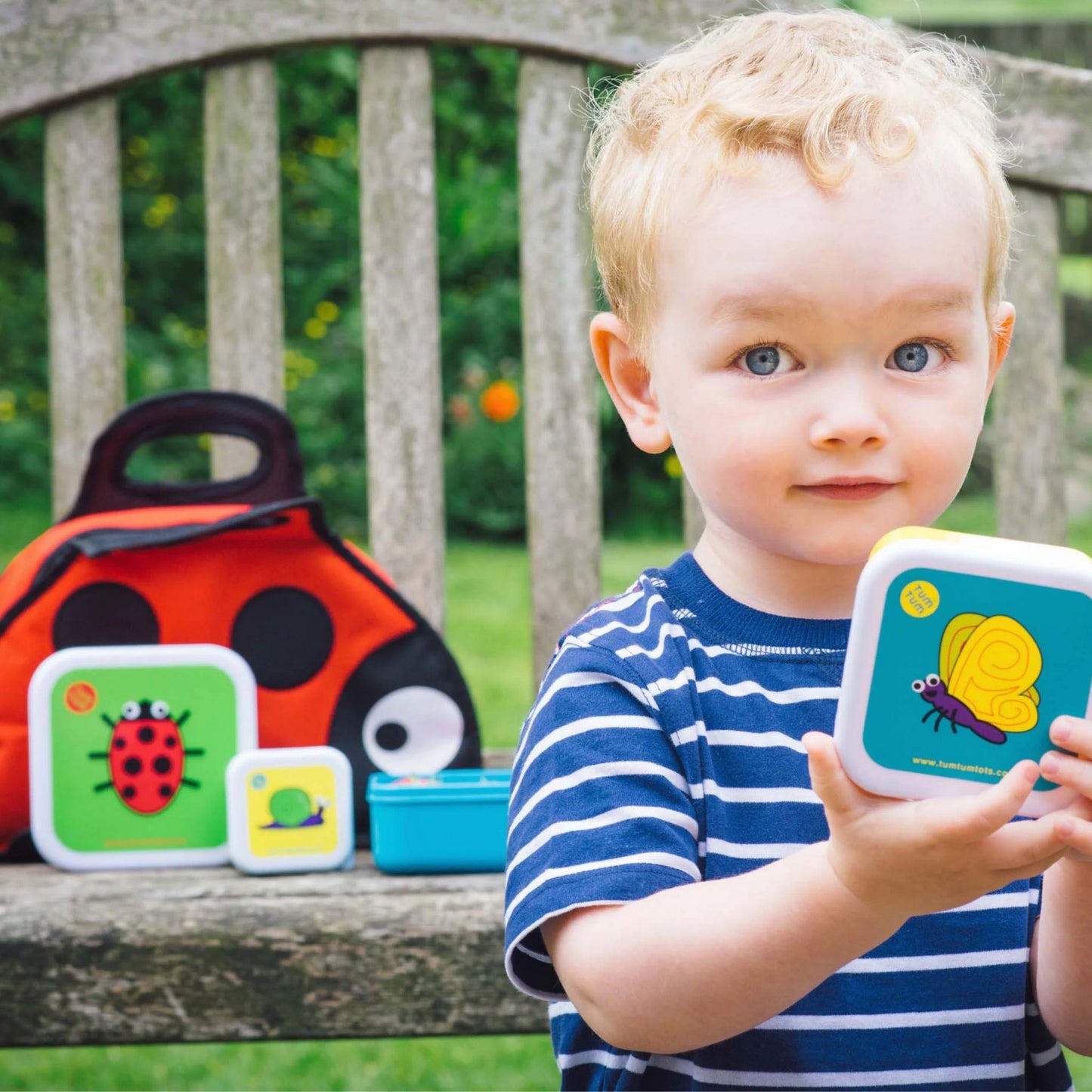 Young child holding a snack pot with colourful bug designs, with a ladybird lunch bag and several other snack pots on a bench in the background.