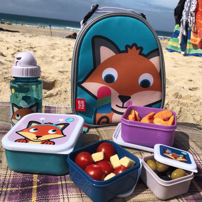 Placed on a beach blanket, a set of snack pots, water bottle, and lunch bag on a beach, filled with snacks like tomatoes, cheese, and peaches.