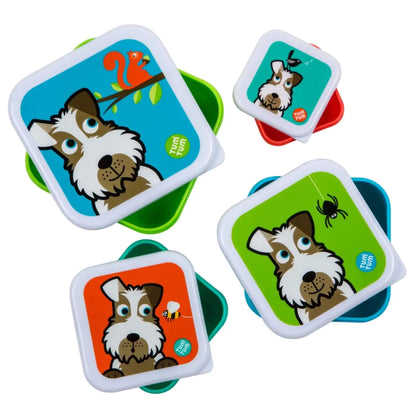 Four square plastic lunch containers with matching lids, each featuring a cartoon dog illustration on a different bright background colour: blue, teal, orange, and green. The containers themselves are translucent green, red, and blue, and the lids have white borders.