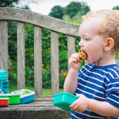 A young child with curly blond hair wearing a blue and white striped shirt is sitting outdoors on a wooden bench, eating a pretzel and holding a green lunchbox. Colourful lunch containers are visible on the bench in the background with a blurred garden setting.