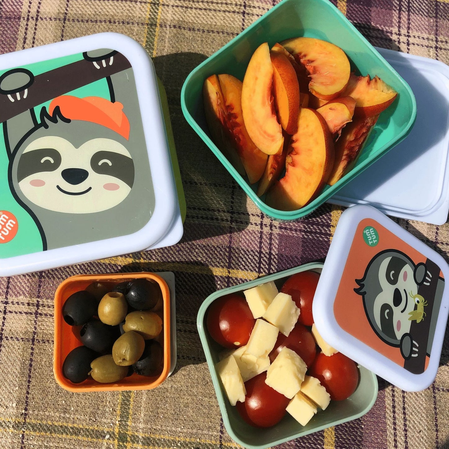 Children's character snack pots with sloth designs, filled with fruit, olives, cheese, and tomatoes, perfect for kids' snacks and lunches. Displayed on a picnic blanket.