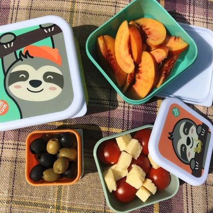 Children's character snack pots with sloth designs, filled with fruit, olives, cheese, and tomatoes, perfect for kids' snacks and lunches. Displayed on a picnic blanket.