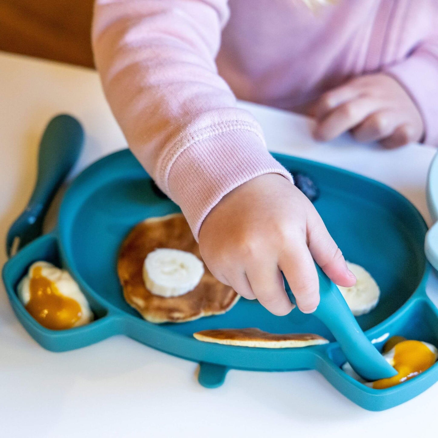 Toddler using blue silicone cutlery and plate to eat pancakes, banana, and yoghurt with fruit, wearing a pink jumper, ideal for self-feeding.
