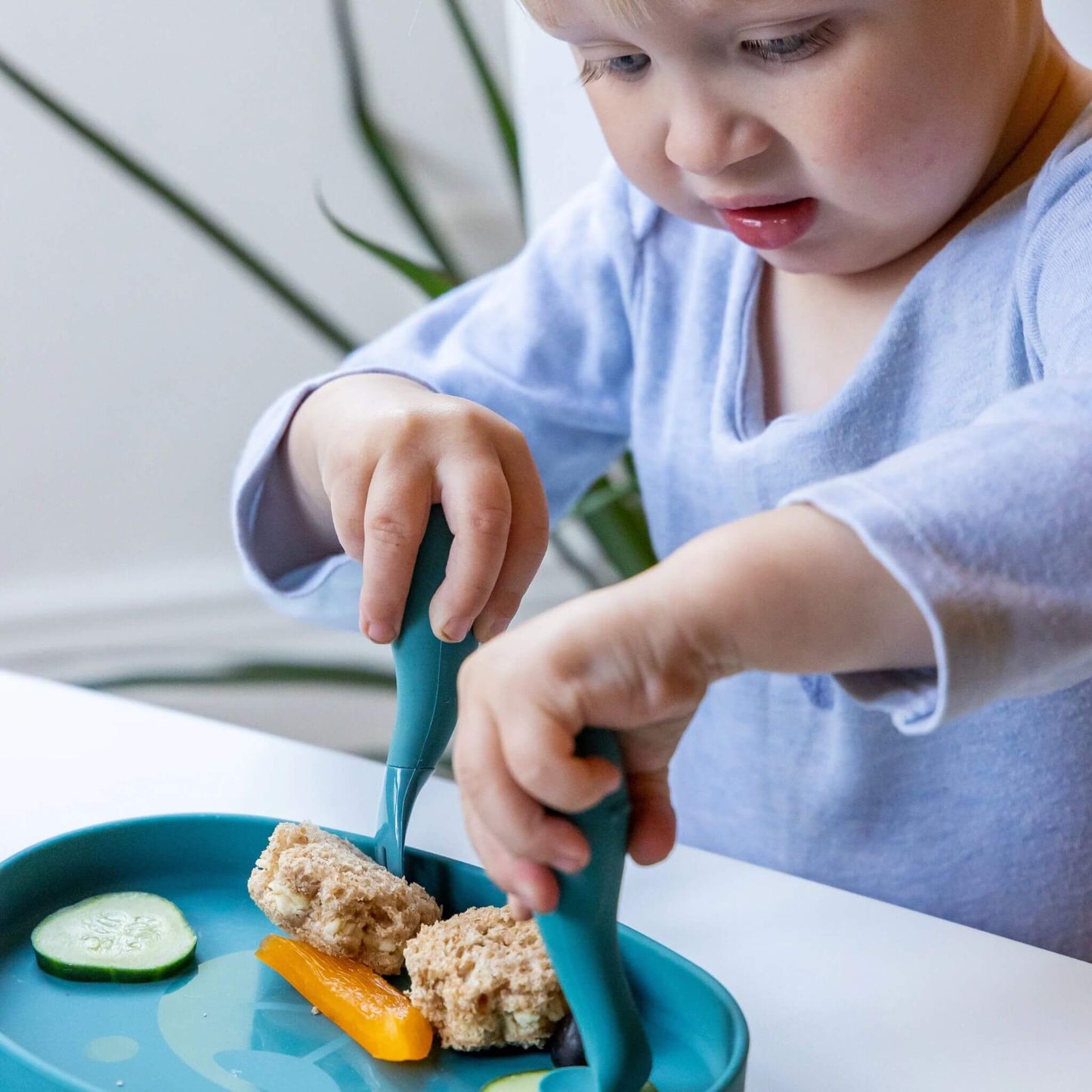 Toddler using blue silicone cutlery with a matching blue plate to eat a sandwich and vegetables, practising self-feeding skills.