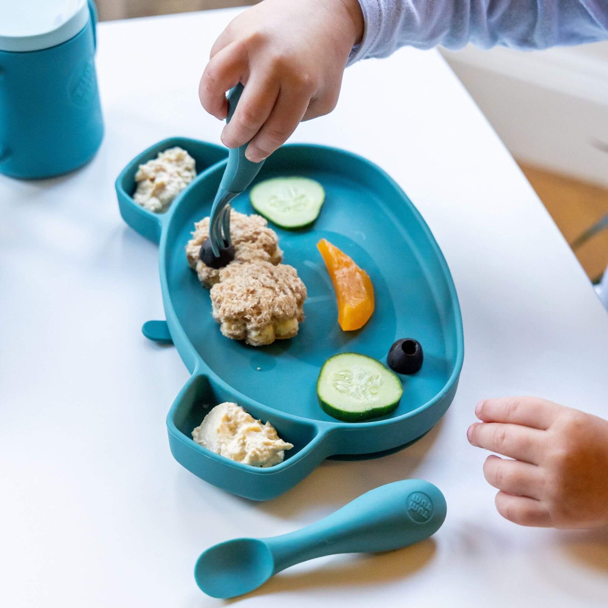Toddler using blue silicone cutlery and plate with cucumber, sandwich, and hummus, ideal for self-feeding, featuring a matching blue cup.
