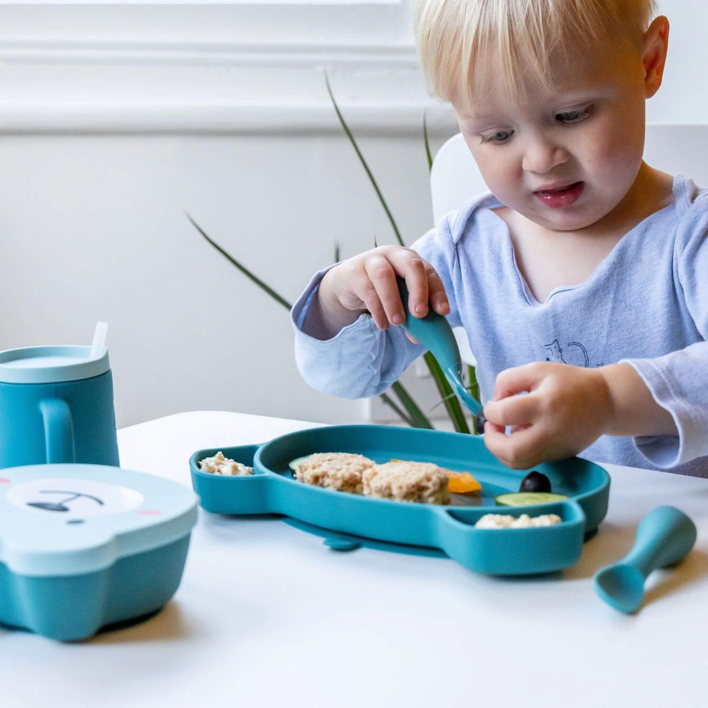 Toddler using blue silicone cutlery with a matching blue plate, cup, and snack box, enjoying a sandwich and vegetables, ideal for self-feeding.