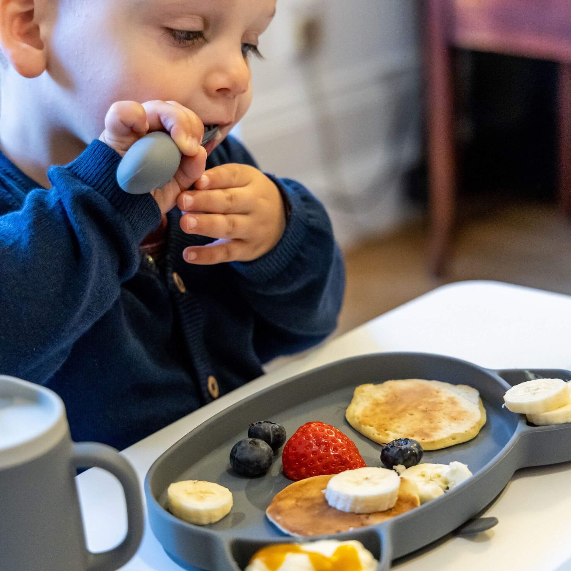 Toddler using silicone cutlery set, enjoying pancakes and fruit on a toddler-friendly plate, perfect for self-feeding.