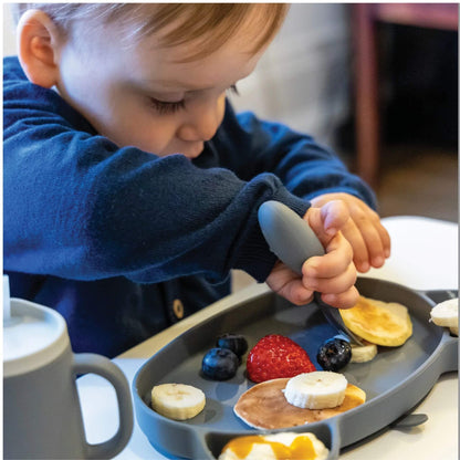 Toddler practising self-feeding with grey silicone cutlery, enjoying pancakes and fruit, promoting independent eating skills.