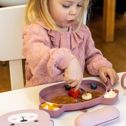 Toddler using pink silicone cutlery and plate with pancakes, banana, and fruit, wearing a pink jumper, ideal for self-feeding.