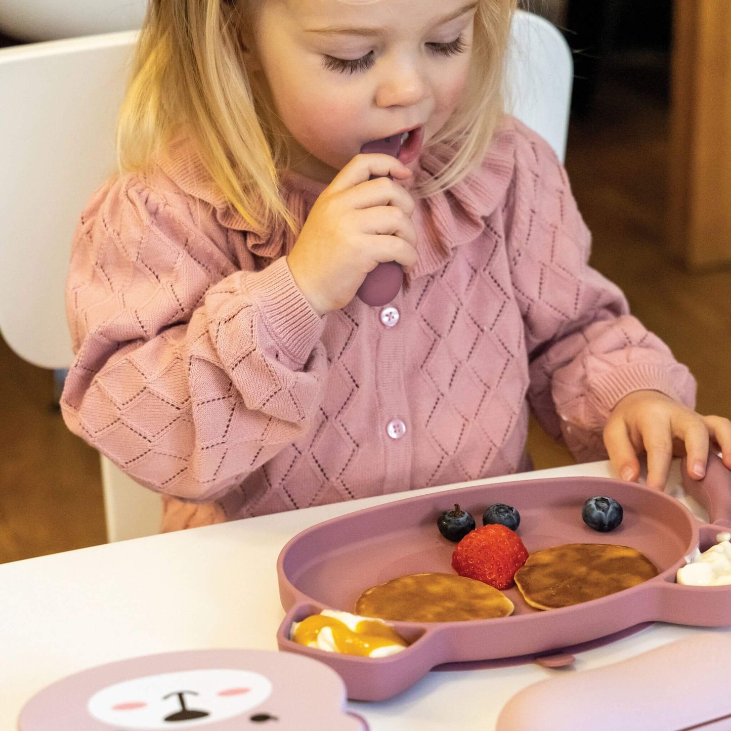 Toddler using pink silicone cutlery and plate with pancakes, fruit, and yoghurt, wearing a pink jumper, perfect for self-feeding.