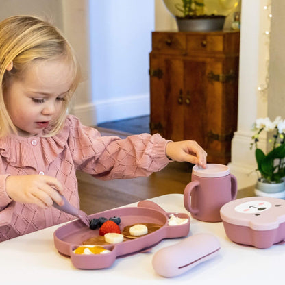 Toddler using pink silicone cutlery, plate, and cup with pancakes, fruit, and yoghurt, wearing a pink jumper, ideal for self-feeding.