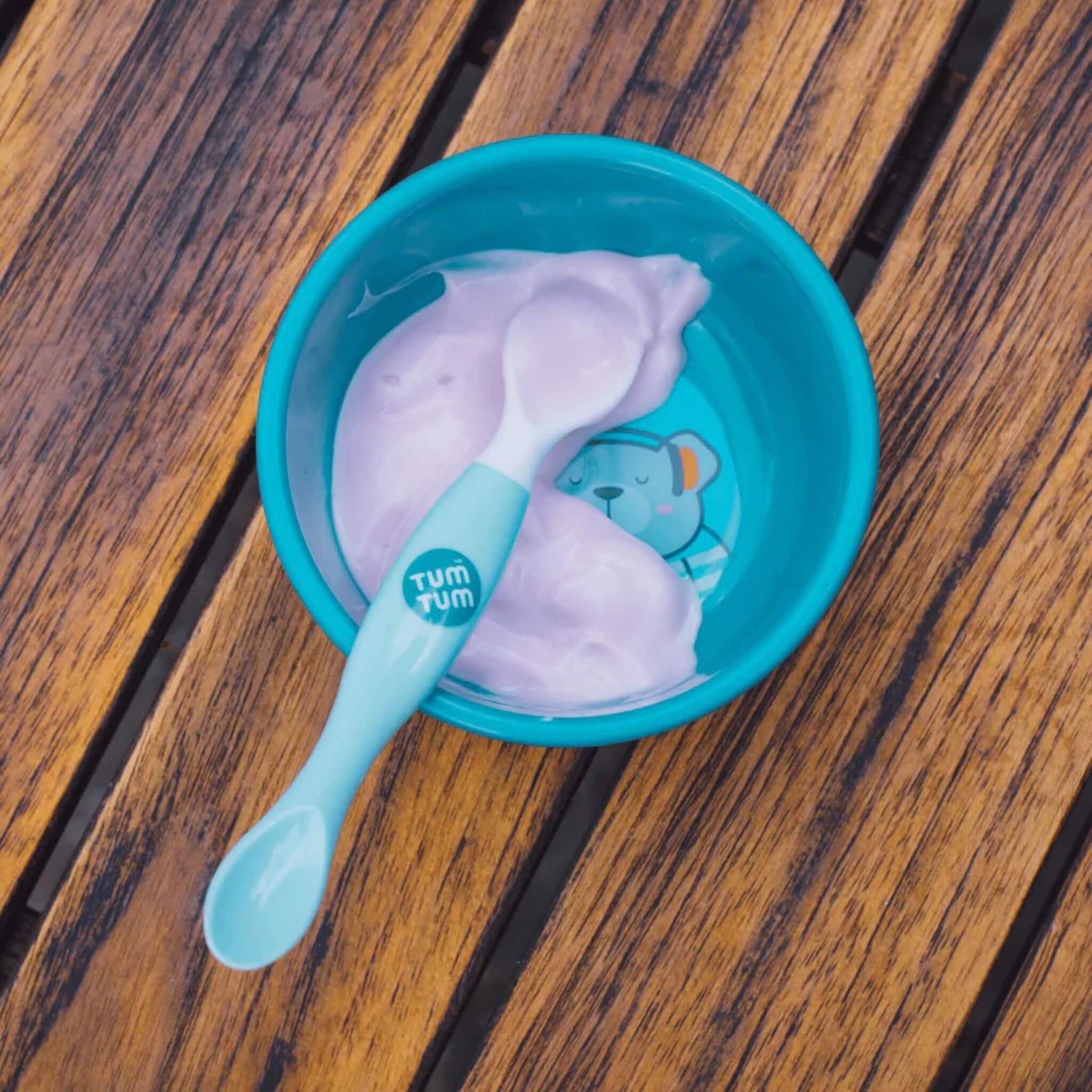 A blue baby bowl containing yoghurt with a blue-handled weaning spoon resting inside, placed on a wooden surface.