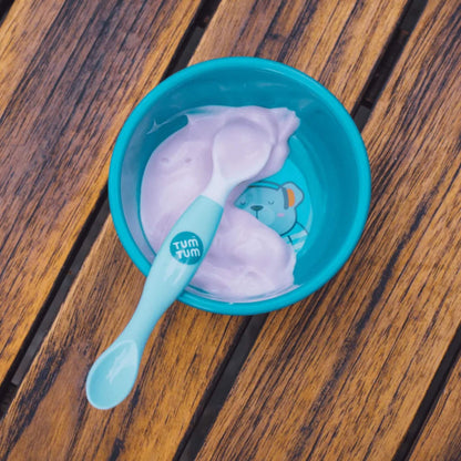 A blue baby bowl containing yoghurt with a blue-handled weaning spoon resting inside, placed on a wooden surface.