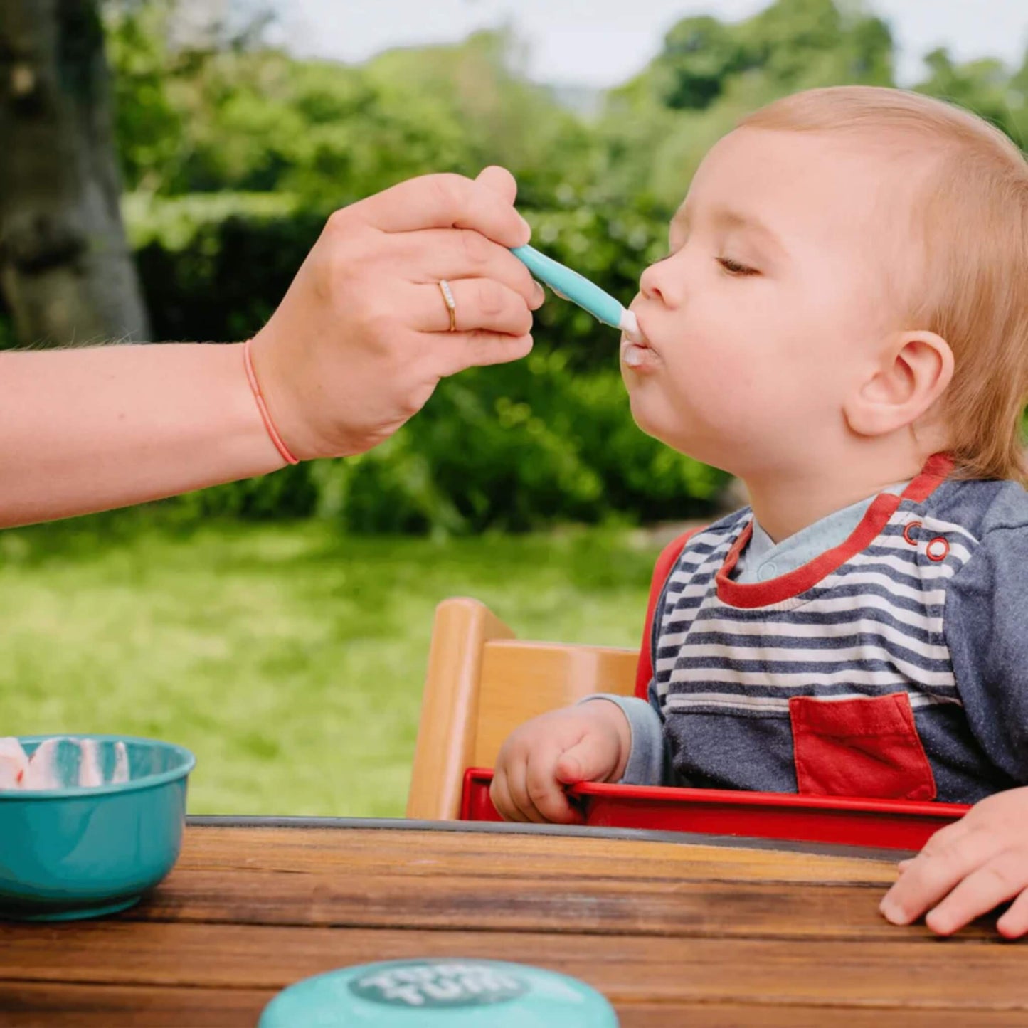 A baby seated in a highchair being fed outdoors with a blue baby spoon, with a matching blue bowl visible on the table.
