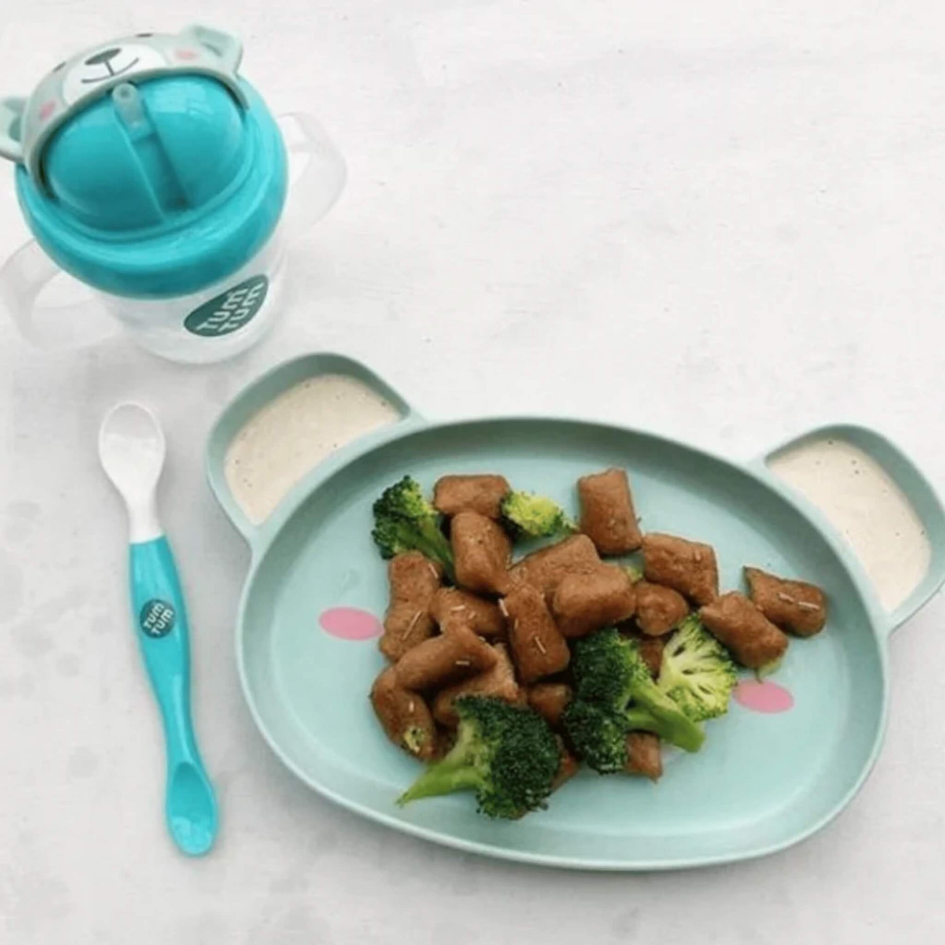 A blue bear-shaped baby plate holding broccoli and finger foods, with a blue spoon and a handled training cup placed beside it.