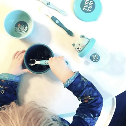 Overhead view of a child using a blue baby spoon to scoop food from a blue bowl, with matching blue feeding accessories arranged on a white table.