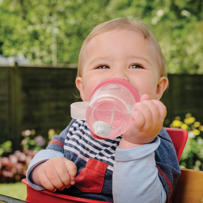 Child holding a pink sippy cup outdoors with greenery in the background
