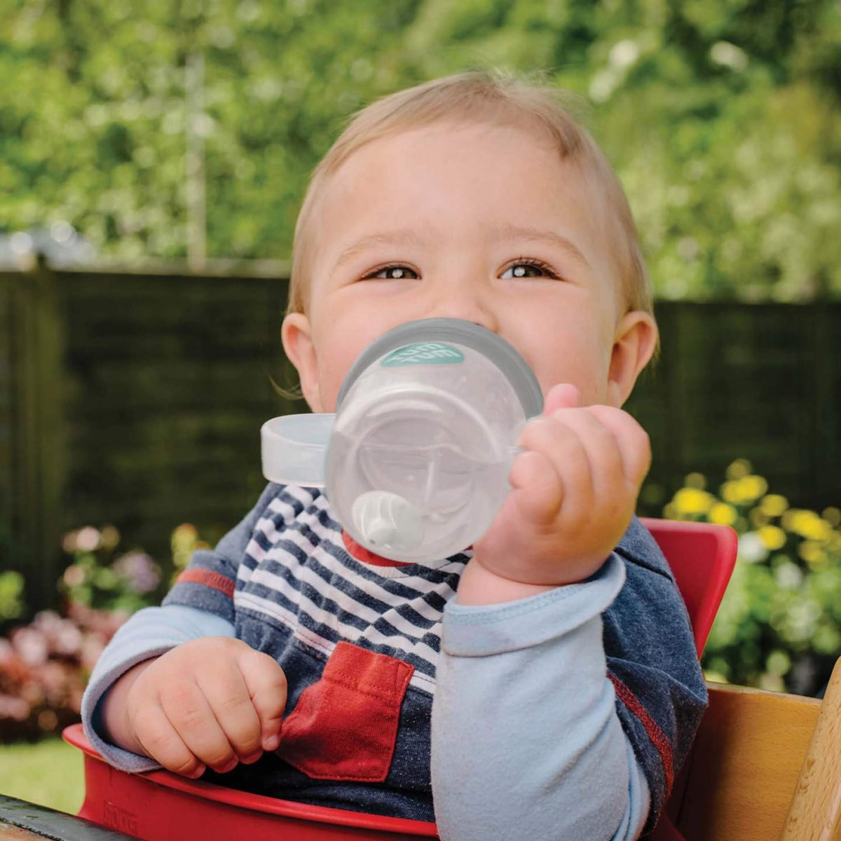 Child holding a sippy cup outdoors with greenery in the background