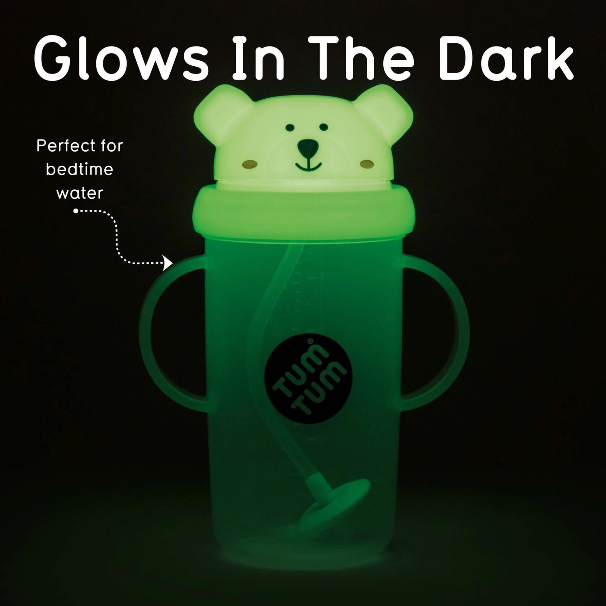 A glow-in-the-dark sippy cup with a bear-shaped lid, shown glowing green in the dark. The cup has two side handles, a straw, and a black circular logo that reads 'tum tum'.