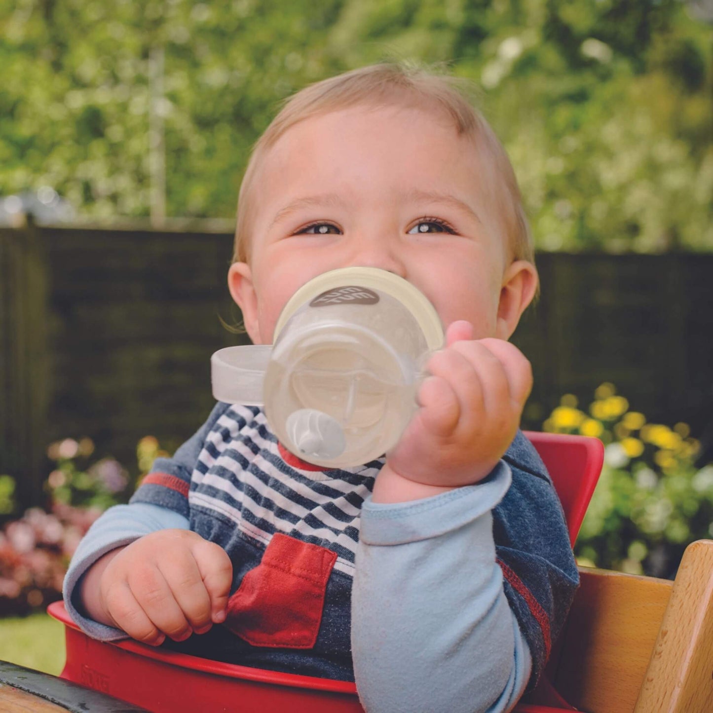 Child holding a sippy cup outdoors with greenery in the background