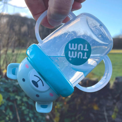 A clear handled straw cup with a blue bear-shaped lid held upside down outdoors, showing the weighted straw hanging down inside the cup to demonstrate how it follows the liquid.