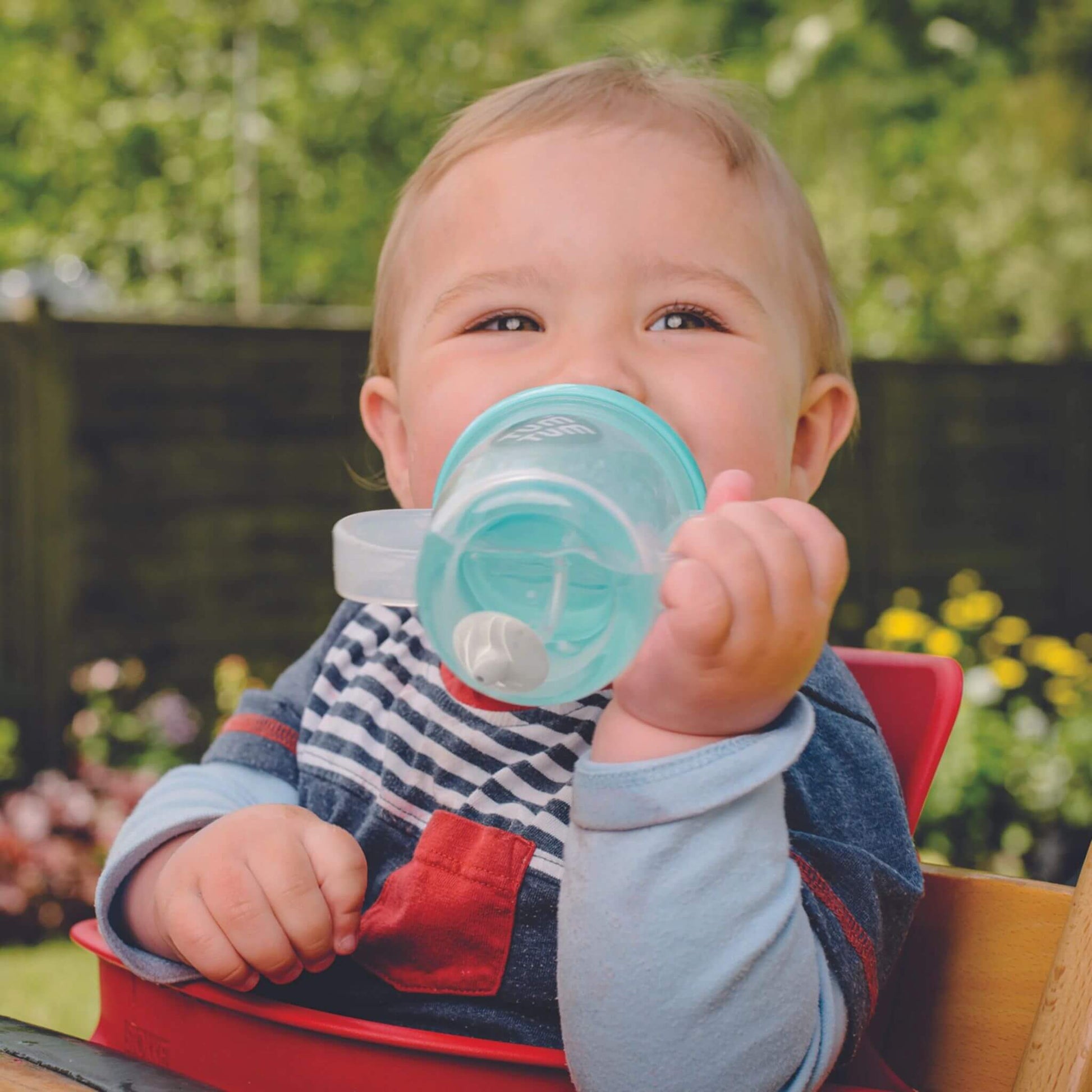 A baby sitting in a highchair holding a clear straw cup with blue lid and side handles, drinking independently while tilting the cup to use the weighted straw.