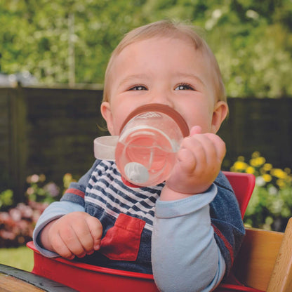 Baby holding a clear handled straw cup with a brown lid and fox character top, drinking while tilting a cup at an angle to show use from different positions.