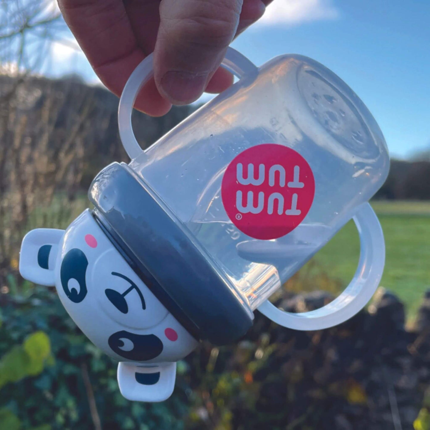 Clear handled cup with white panda character lid shown tilted sideways outdoors, demonstrating weighted straw following liquid inside cup, with grey lid and transparent handles visible.