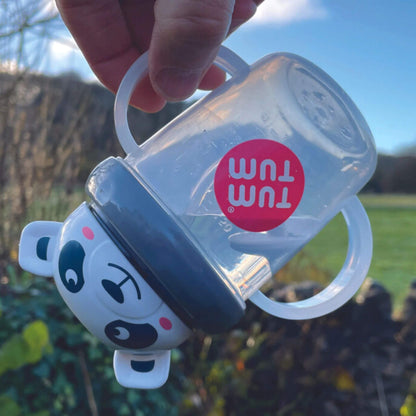 Clear handled cup with white panda character lid shown tilted sideways outdoors, demonstrating weighted straw following liquid inside cup, with grey lid and transparent handles visible.