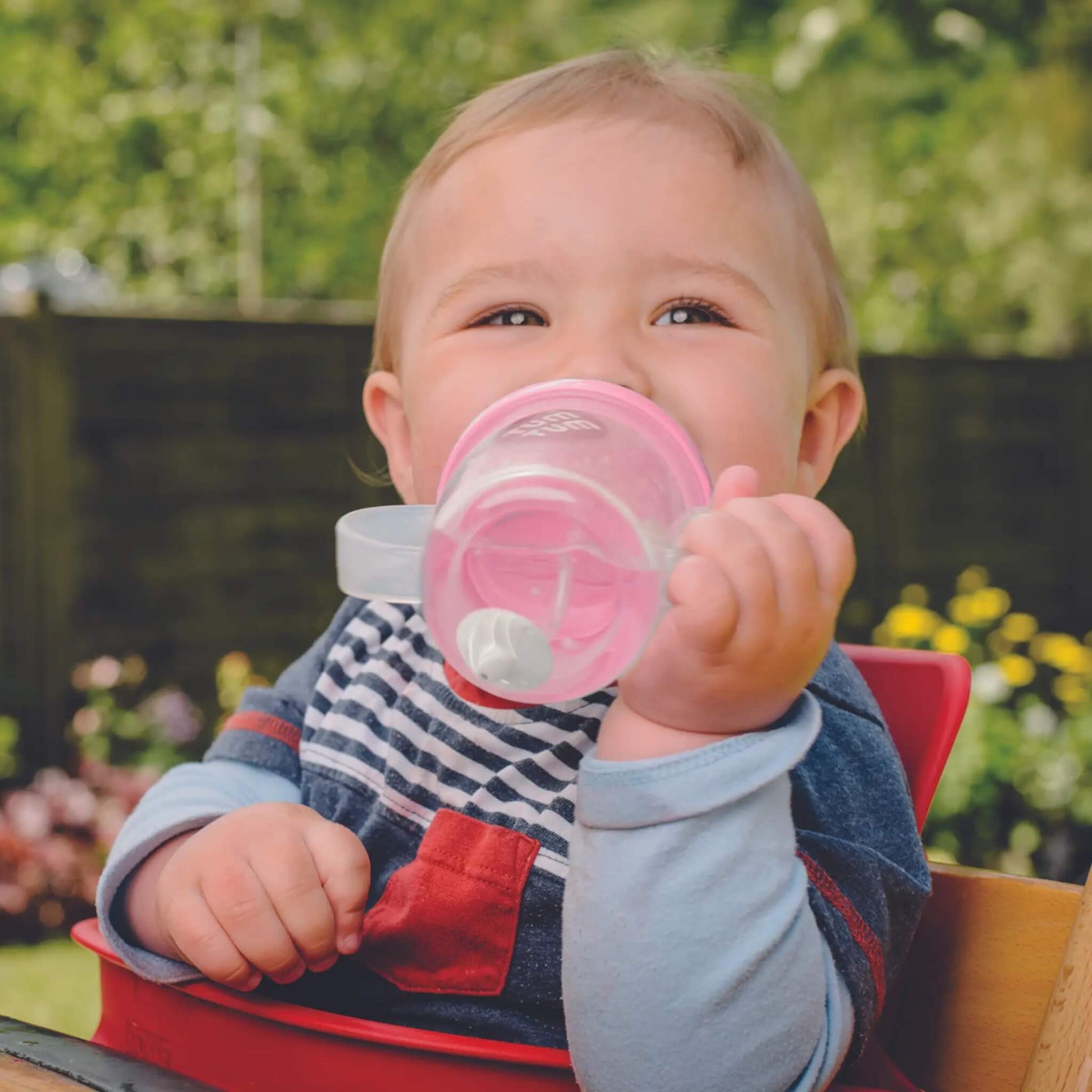Baby holding a pink sippy cup outdoors with greenery in the background