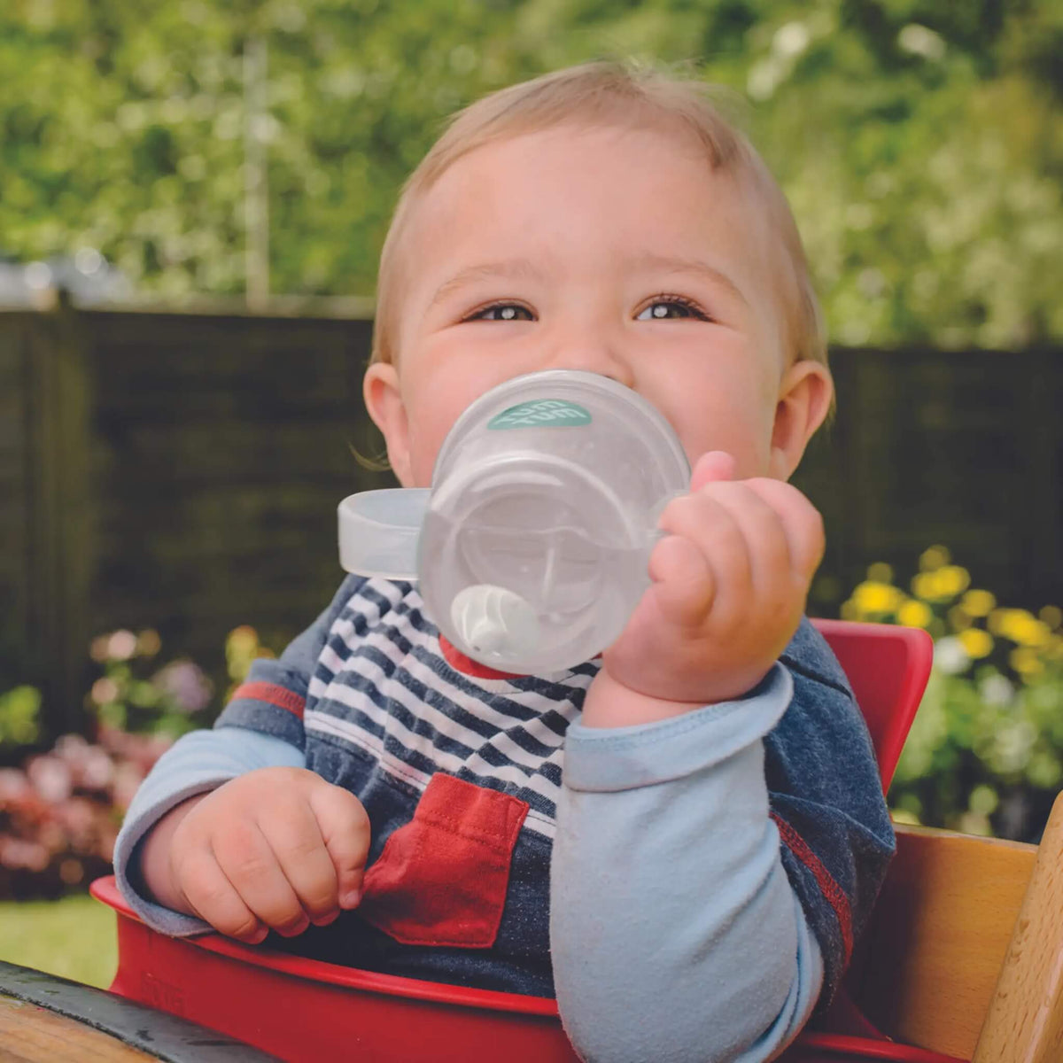 Child holding a sippy cup outdoors with greenery in the background