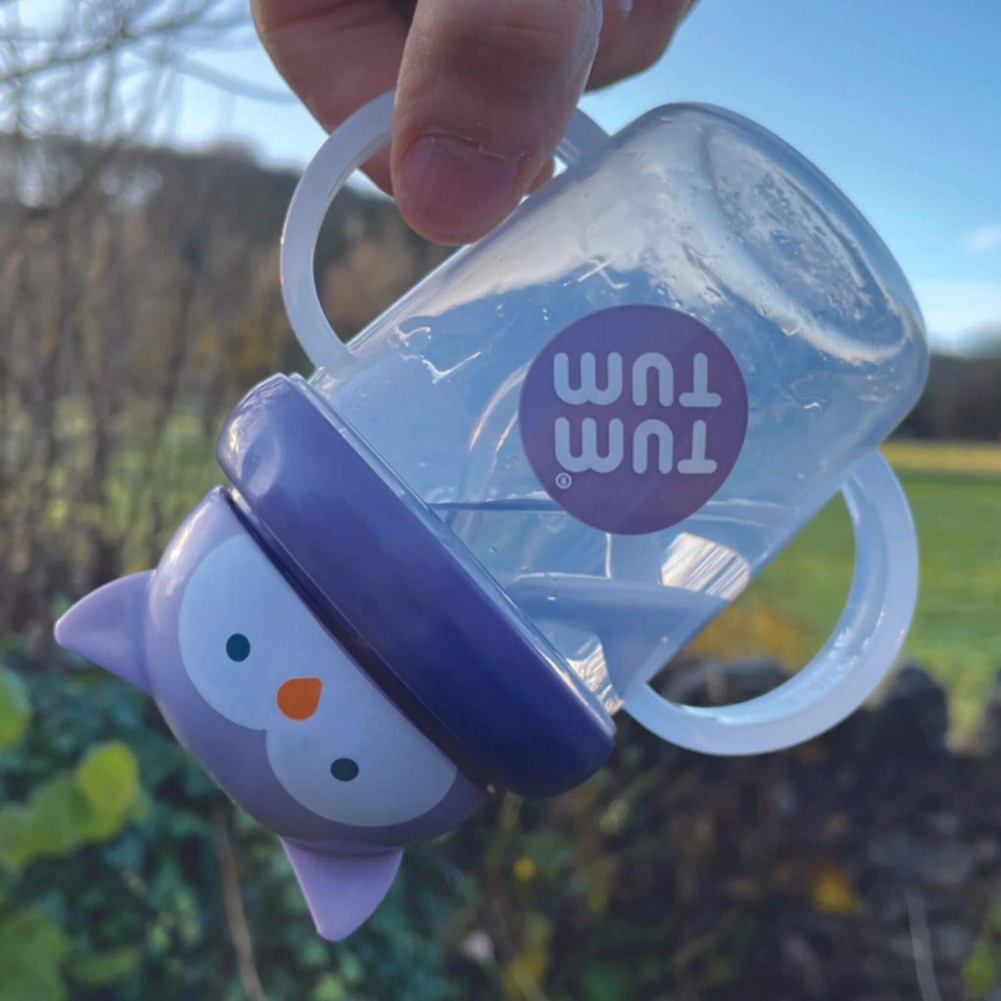 Clear handled cup with purple owl lid tilted upside down outdoors, showing water flowing inside as a weighted straw follows liquid for drinking at any angle.