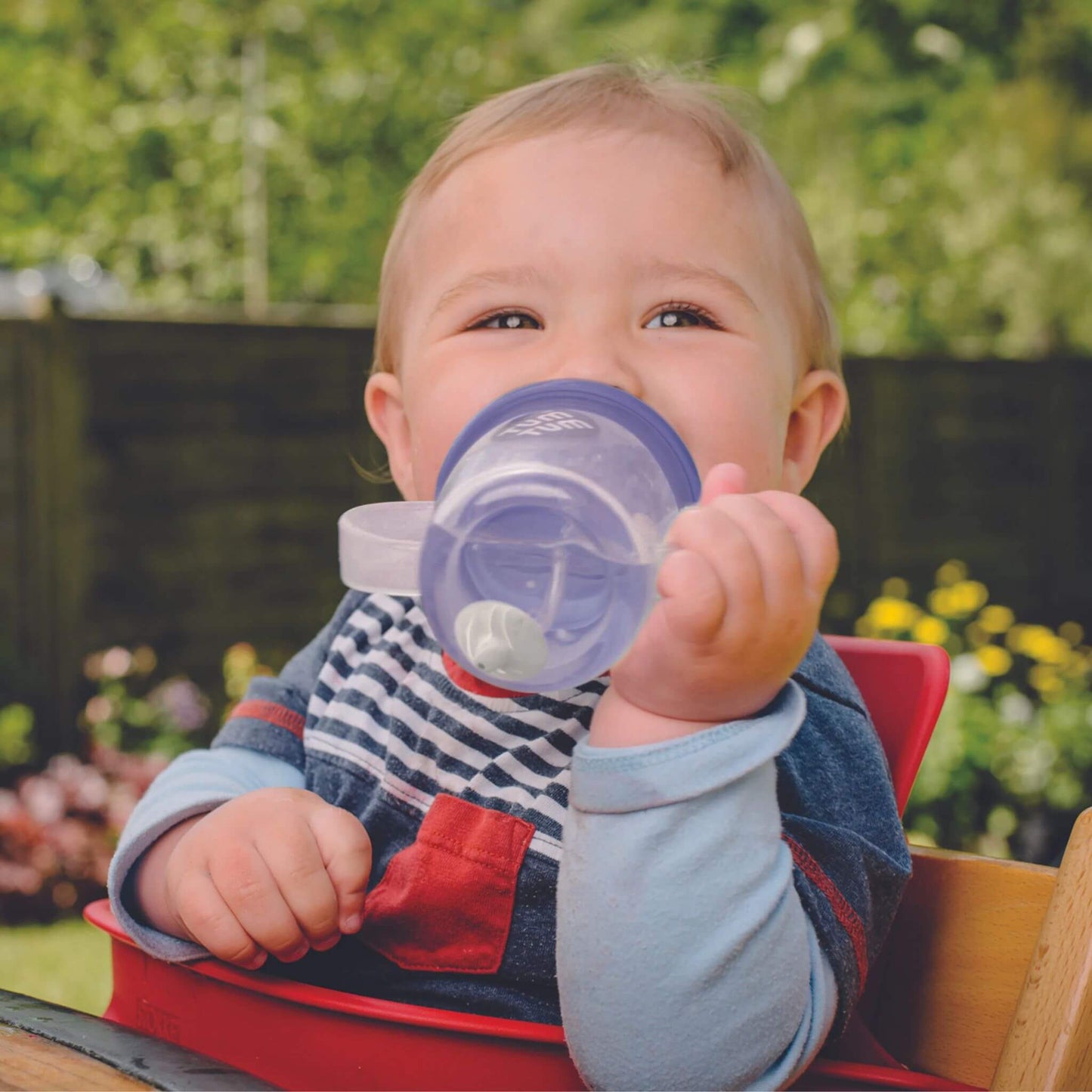 Baby sitting outdoors holding a clear handled cup with a purple lid, drinking independently while gripping the cup with one hand.