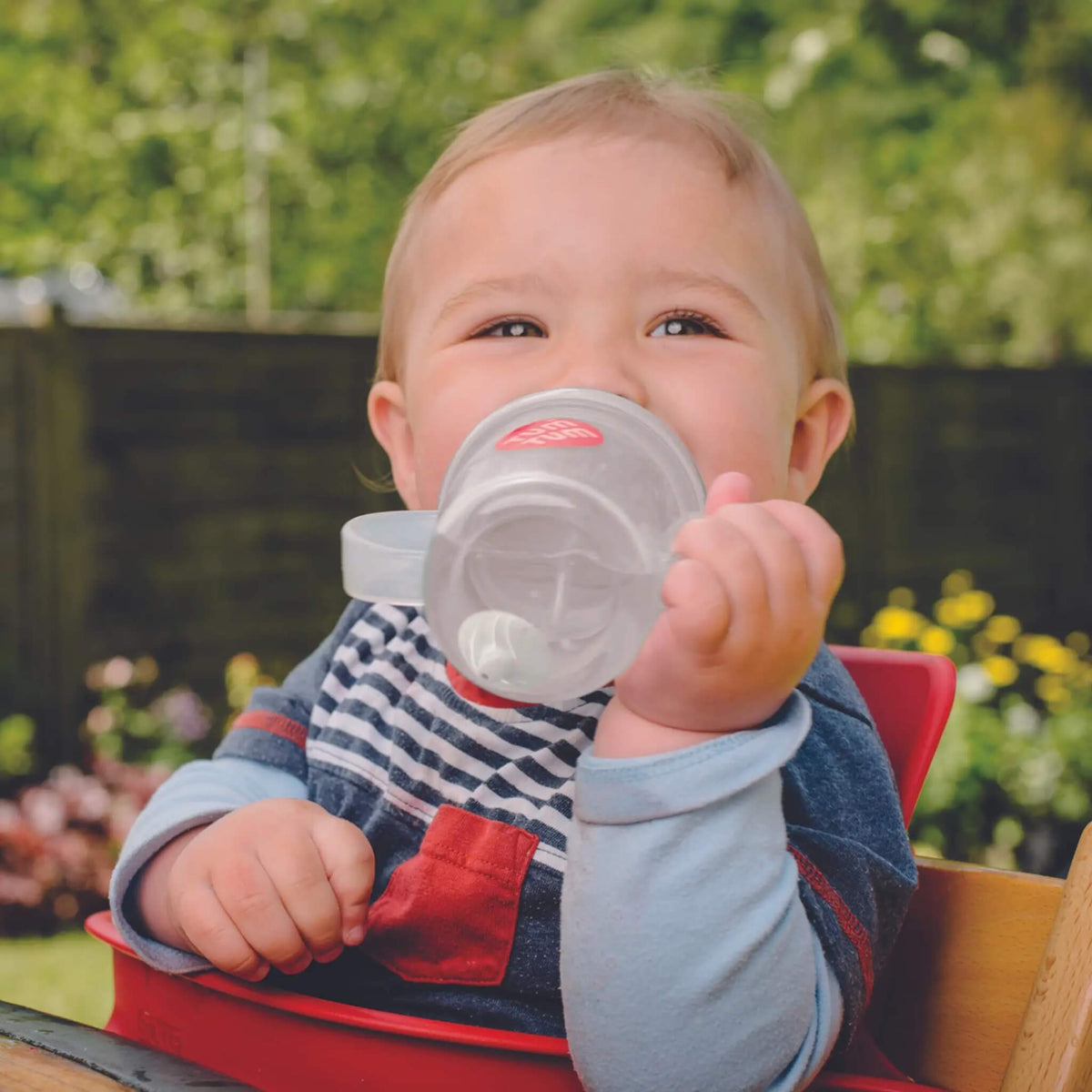 A baby sitting in a red high chair outdoors, holding a clear plastic sippy cup with a white lid near their mouth. The baby is wearing a blue and white striped bib over a light blue long-sleeve shirt.