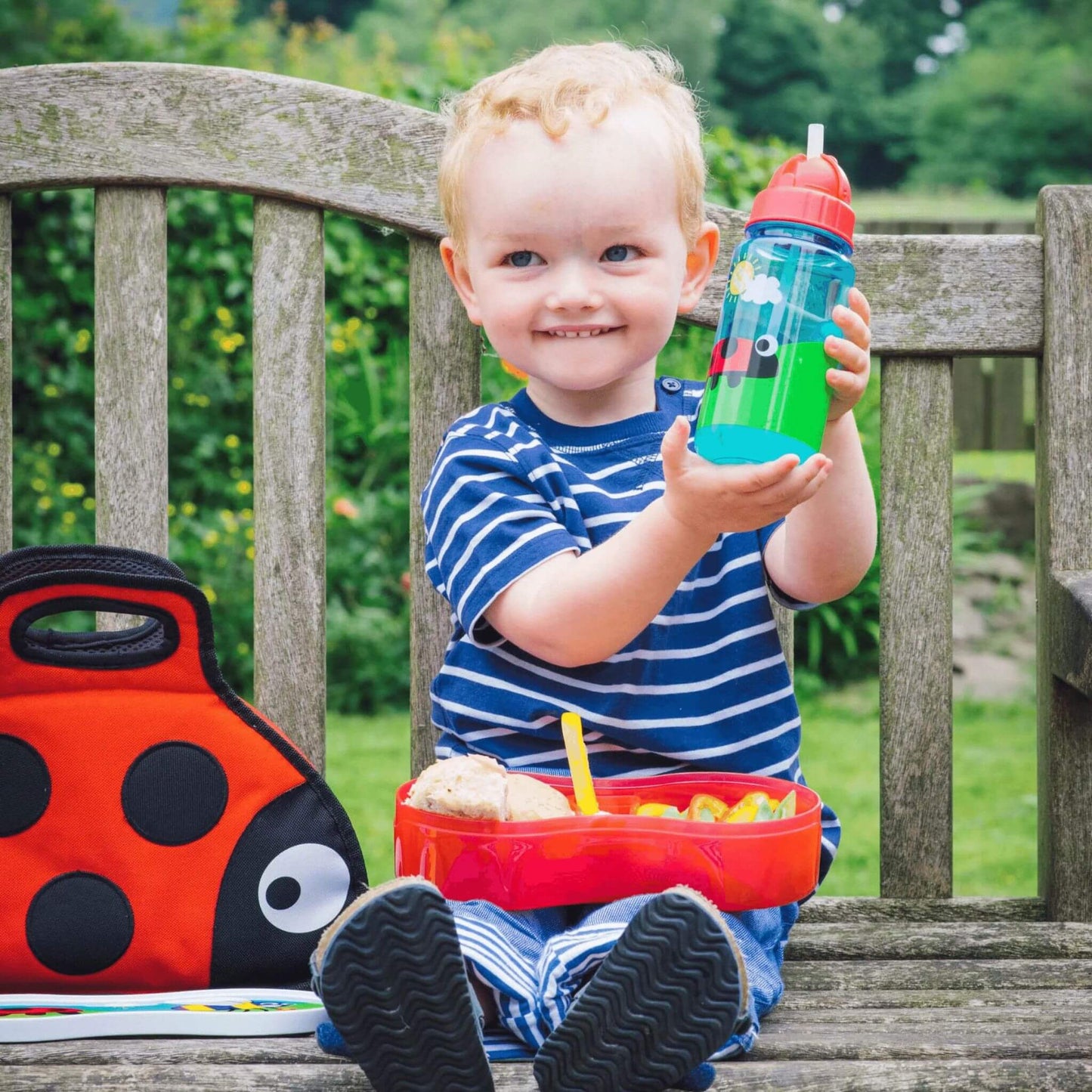 Smiling child holding a brightly illustrated water bottle, sitting with matching ladybird lunchbox and bag, perfect for kids on the go.