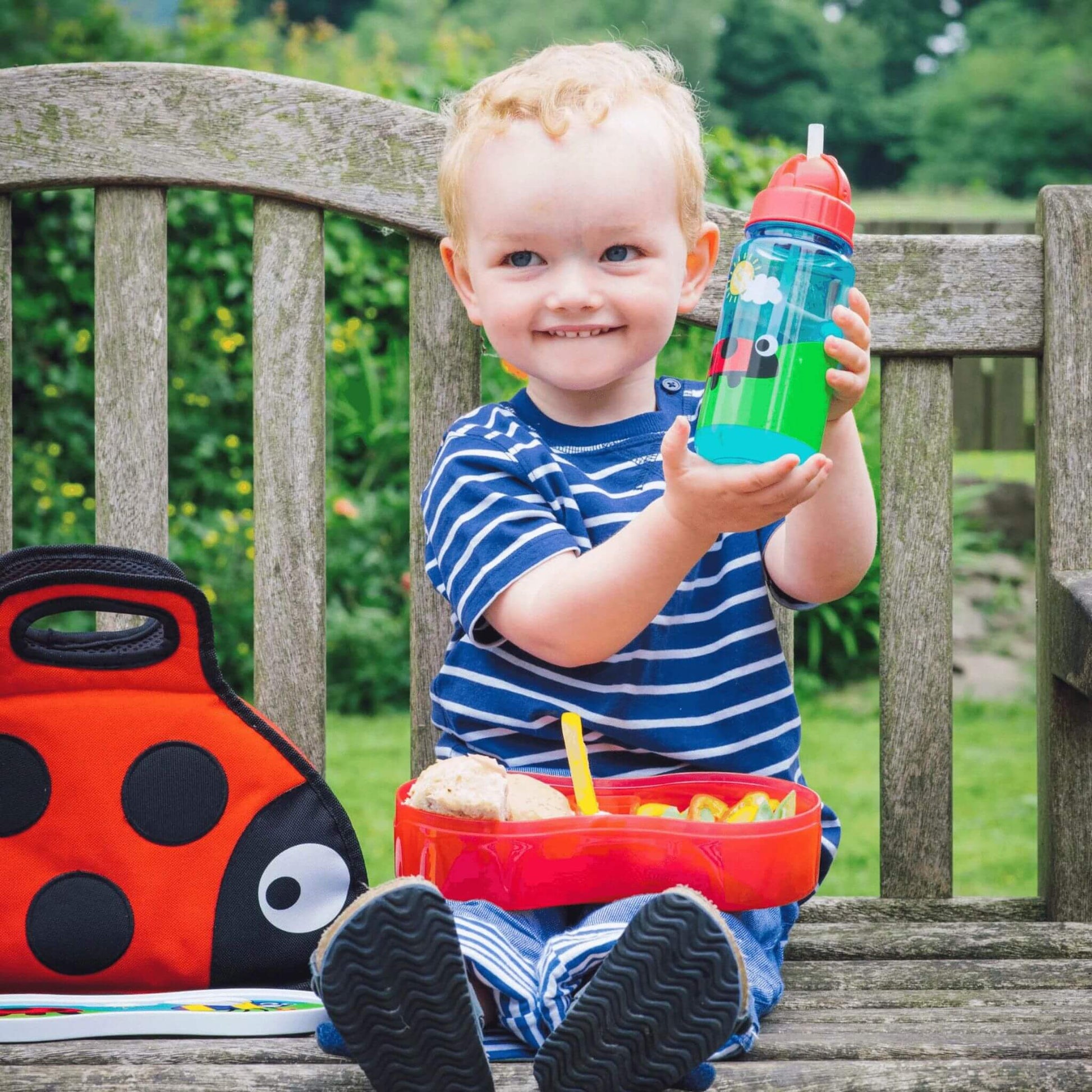 Smiling child holding a brightly illustrated water bottle, sitting with matching ladybird lunchbox and bag, perfect for kids on the go.