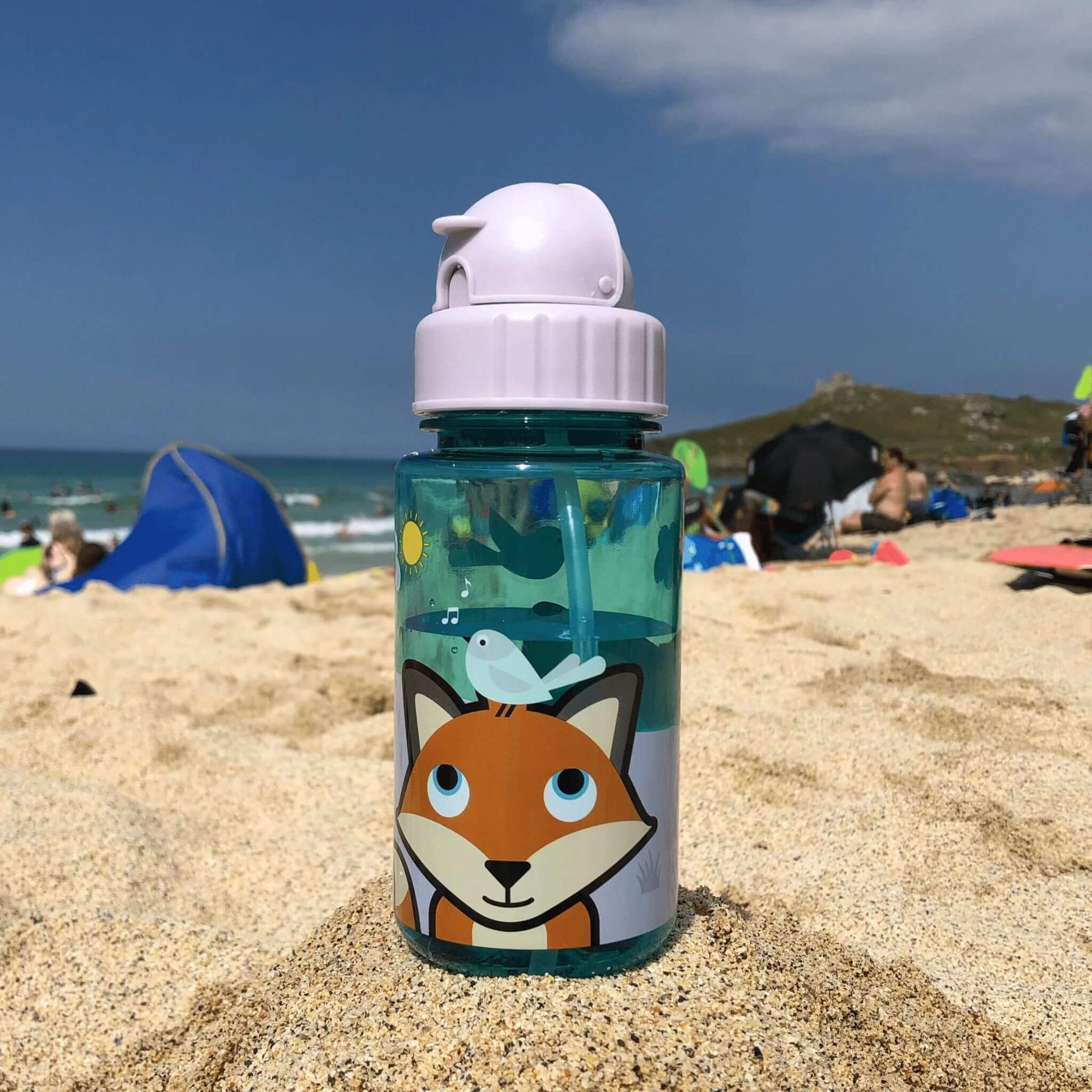 A toddler water bottle with Felicity Fox design, featuring a flip top lid and built-in straw, placed on a sandy beach with people in the background.