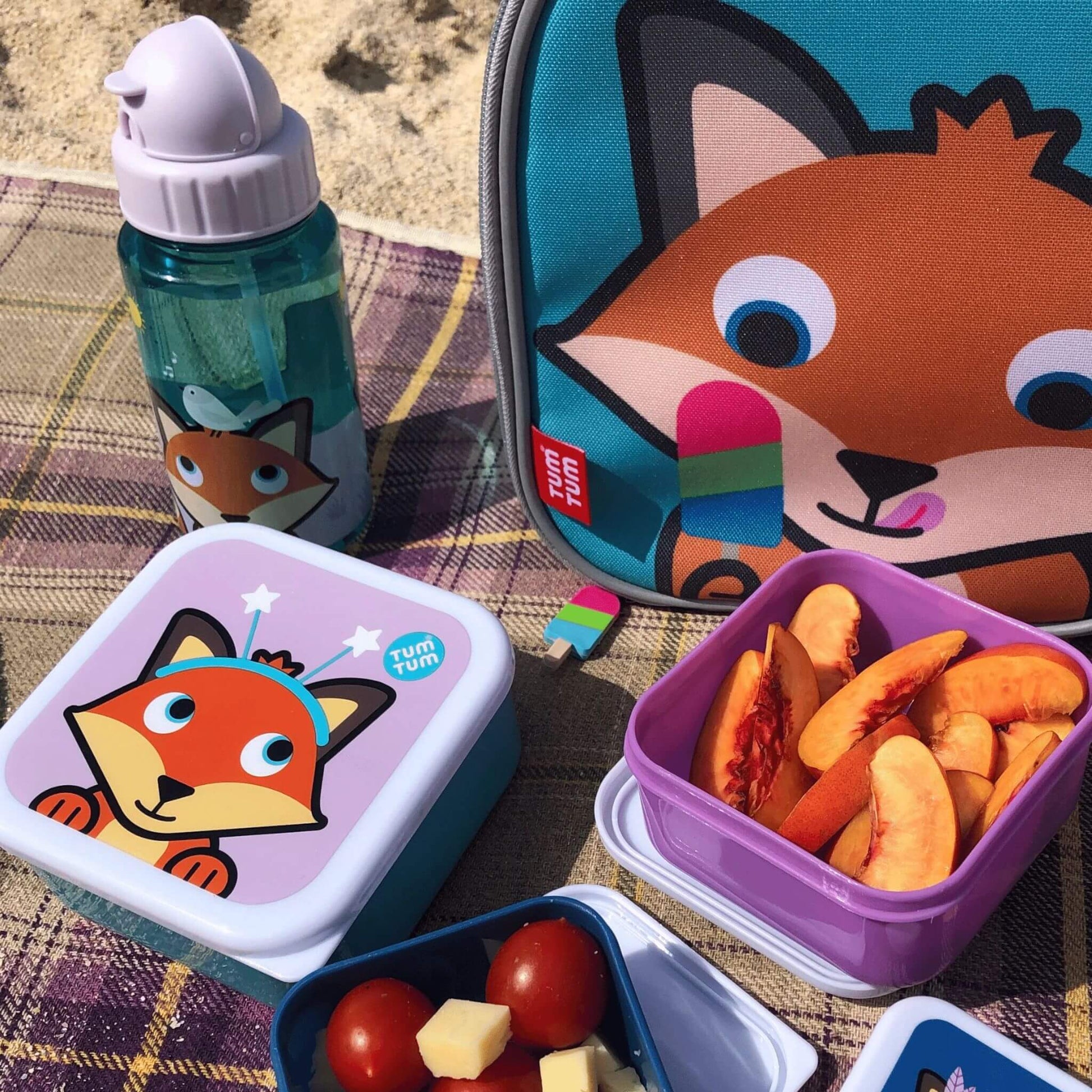 Kid's water bottle with Felicity Fox character design, alongside matching snack boxes and lunch bag, placed on a blanket at the beach, perfect for toddler meals.