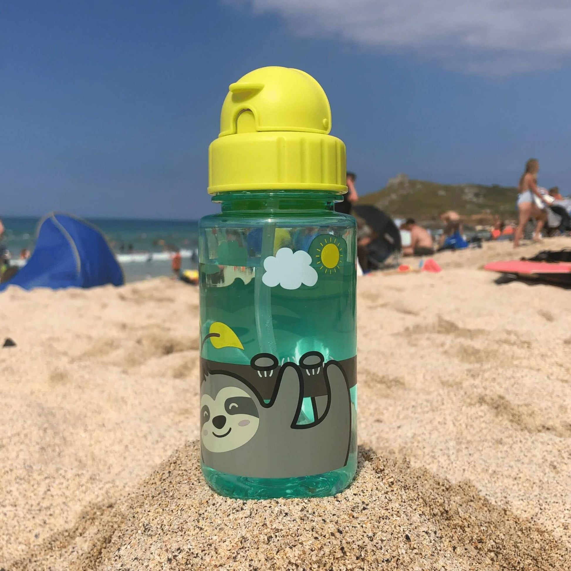 Kid's water bottle with sloth design, featuring a flip top lid and built-in straw, placed on a sandy beach with people in the background.