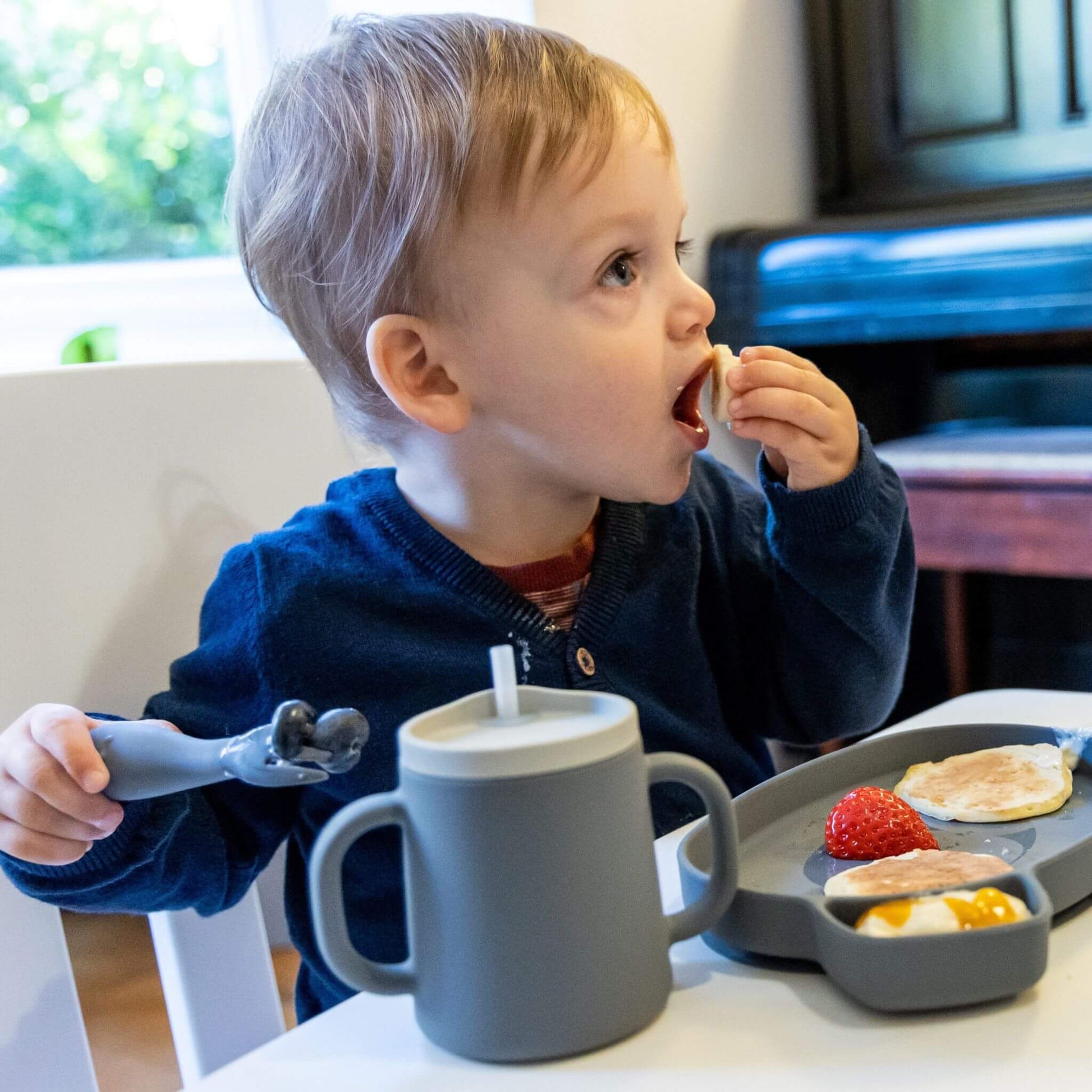 Toddler using  grey TUM TUM silicone cutlery and  cup, enjoying a meal of pancakes, fruit, and yoghurt on a plate, ideal for self-feeding.