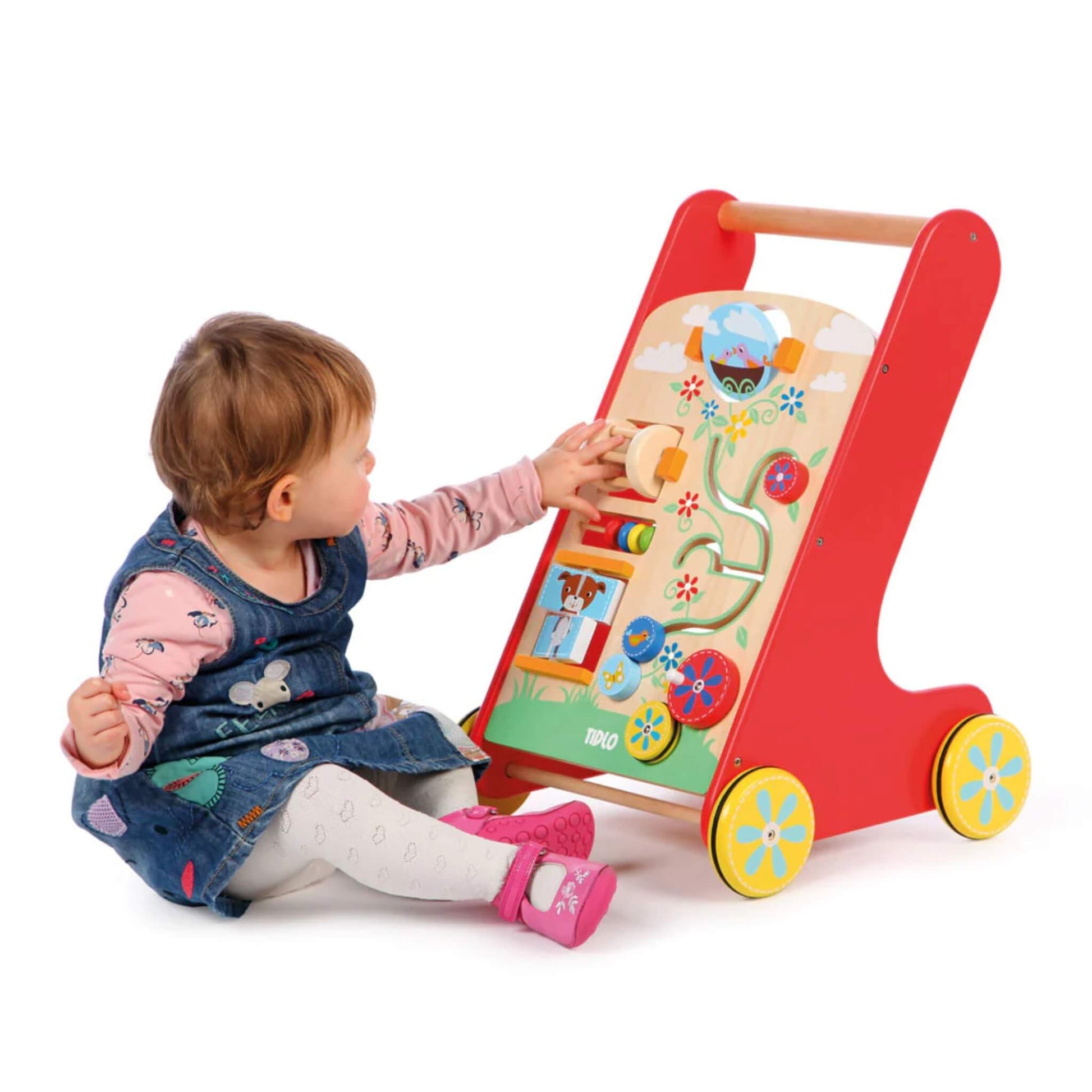 A toddler in a denim dress interacts with the Tidlo Activity Walker, seated beside it and reaching for the spinning activity pieces on the wooden front panel.