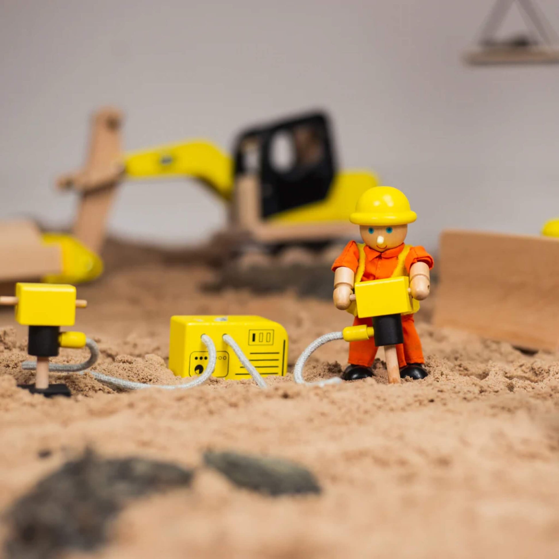 Close-up of a wooden construction figure using the jackhammer from the Tidlo Construction Equipment Set in a sand play setup with a digger in the background.